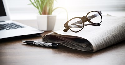 Pöydällä on sanomalehti, kannettava tietokone, silmälasit ja kuulakärkikynä.