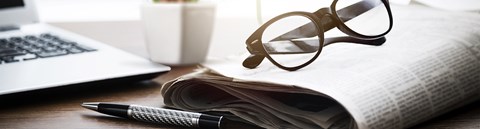 On the table is a newspaper, a laptop, glasses and a pen.