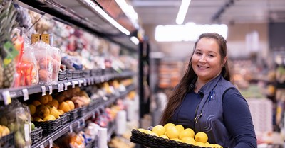 A woman looks at the camera in the refrigerated section of the grocery store. He is smiling and has a fruit basket on his lap.