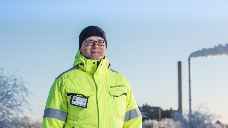 Työtakkiin pukeutunut Oulun Energian työntekijä hymyilee kameralle. Taustalla Toppilan voimalaitos.