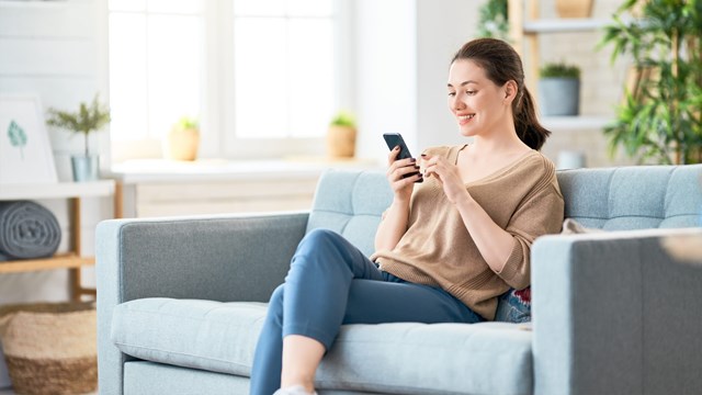 A smiling woman sits on the couch with her smartphone in her hand.