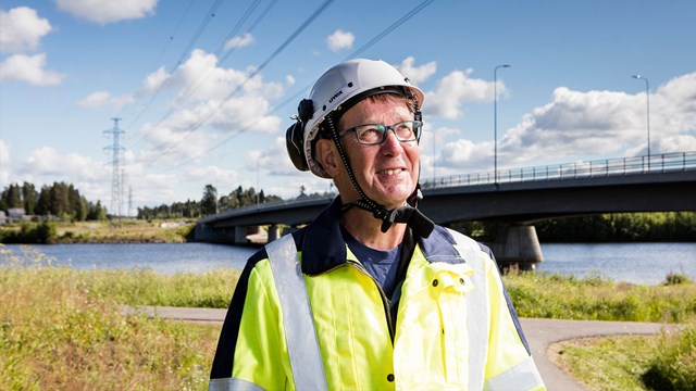 Oulun Energian työntekijä katsoo hymyillen kaukaisuuteen. Taustalla joki ja voimalinja.